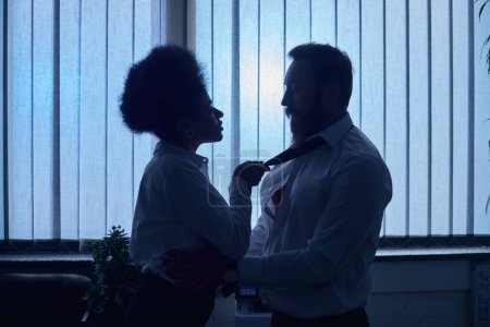 Téléchargez les photos : Silhouette sombre de femme afro-américaine tirant cravate de collègue et le séduisant dans le bureau - en image libre de droit
