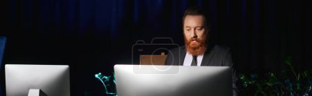 Foto de Trabajo de horas extras, hombre de negocios barbudo concentrado de pie monitor de computadora en la oficina por la noche, pancarta - Imagen libre de derechos