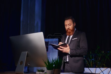 Téléchargez les photos : Élégant homme d'affaires barbu écrit dans un ordinateur portable près de moniteur d'ordinateur la nuit au bureau, de longues heures - en image libre de droit