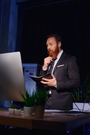 Téléchargez les photos : Homme d'affaires barbu réfléchi avec ordinateur portable regardant moniteur d'ordinateur et travaillant dans le bureau de nuit - en image libre de droit