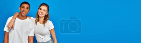 happy young woman embracing african american friend on blue backdrop, cultural diversity, banner