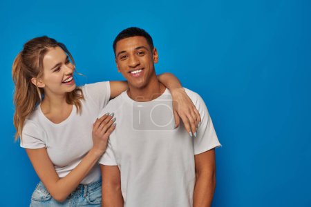 Photo for Happy young woman embracing african american friend on blue backdrop, positive emotion and diversity - Royalty Free Image