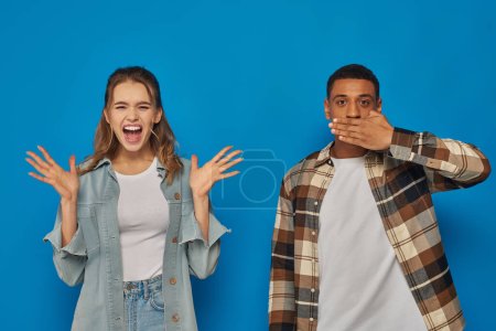 Reaktion eines interrassischen Paares auf blauem Hintergrund, emotionale Frau schreit in der Nähe eines afrikanisch-amerikanischen Mannes