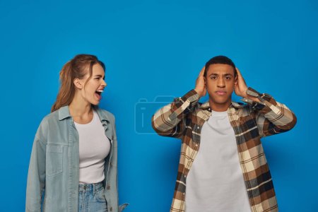mujer emocional gritando cerca de hombre afroamericano cubriendo sus oídos sobre fondo azul, expresivo