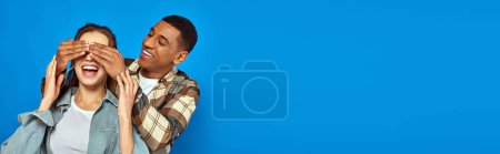 excited african american man covering eyes of woman with open mouth on blue background, banner