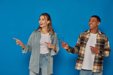 Photo for Happy woman with open mouth and african american man pointing away with fingers on blue background - Royalty Free Image