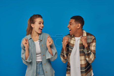 happy interracial couple screaming from joy looking at each other on blue backdrop, emotional people