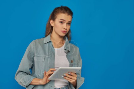 schöne junge Frau mit digitalem Tablet und Blick auf Kamera auf blauem Hintergrund, verwirrtes Gesicht