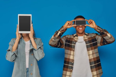 Téléchargez les photos : Couple interracial tenant smartphone et tablette numérique avec écran blanc sur fond bleu - en image libre de droit