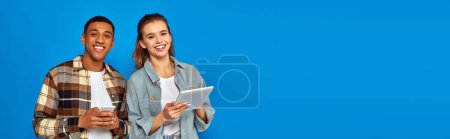 happy interracial couple holding smartphone and digital tablet on blue backdrop, banner