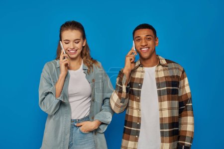 Cheerful woman and happy african american man having phone call, using smartphones on blue backdrop