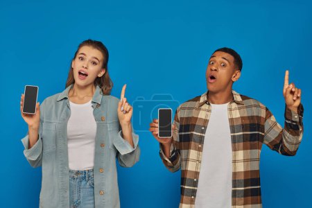 cheerful woman and happy african american holding smartphones and pointing up on blue backdrop
