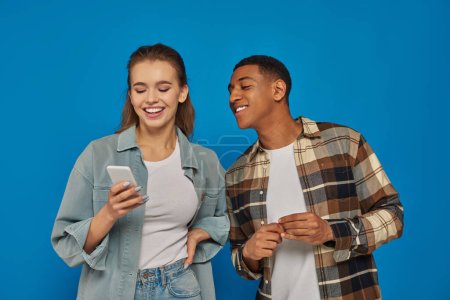 happy african american man looking at  smartphone of female friend and smiling on blue backdrop