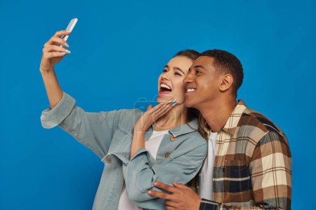 alegre pareja interracial tomando selfie y sonriendo mientras mira el teléfono inteligente en azul telón de fondo