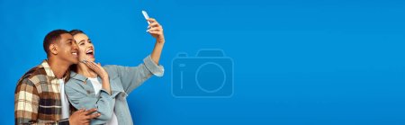 cheerful interracial couple taking selfie while looking at smartphone on blue backdrop, banner