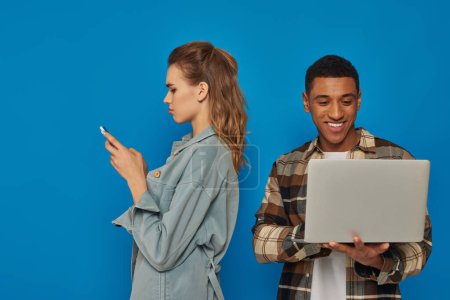 happy african american freelancer using laptop near sad woman chatting on smartphone, blue backdrop