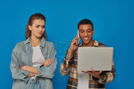 femme ennuyée regardant l'homme afro-américain souriant et utilisant un ordinateur portable, ayant un appel téléphonique sur bleu