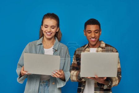 happy interracial freelancers using laptops on blue backdrop, diverse cultures man and woman