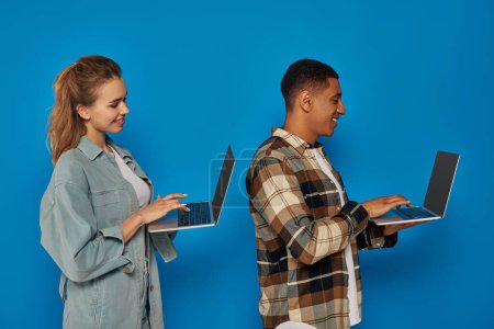 Téléchargez les photos : Heureux interracial pigistes en utilisant des ordinateurs portables sur fond bleu, diverses cultures homme et femme, côté - en image libre de droit