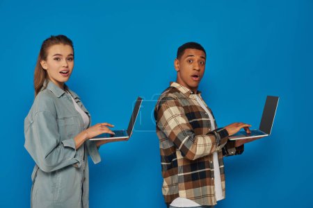 amazed  interracial freelancers using laptops on blue backdrop, diverse cultures man and woman