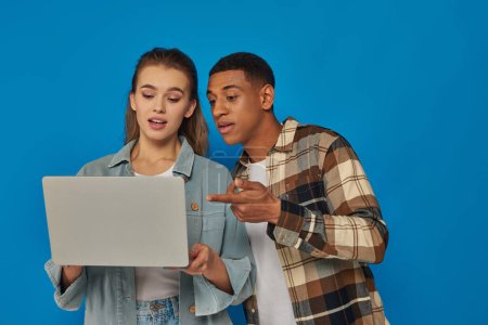 Téléchargez les photos : Homme afro-américain pointant vers ordinateur portable et discuter des idées avec une collègue féminine sur fond bleu - en image libre de droit