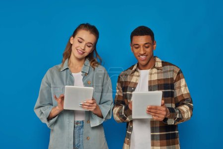happy interracial man and woman using digital tablets on blue backdrop, social media and networking