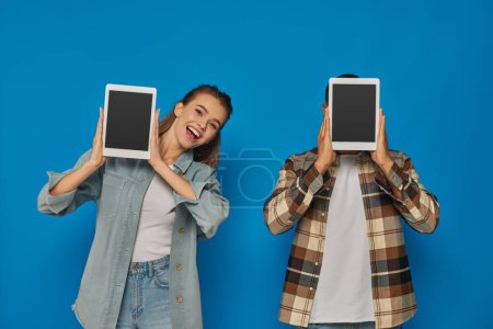 Afrikanisch-amerikanischer Mann verschleiert Gesicht mit digitalem Tablet nahe aufgeregter Frau auf blauem Hintergrund