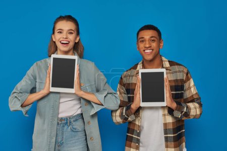 happy interracial man and woman holding digital tablets with blank screen on blue backdrop