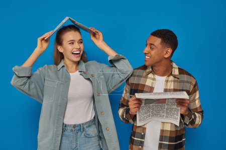 happy woman covering head with newspaper near smiling african american man on blue backdrop