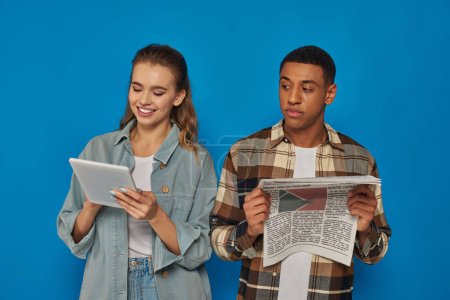 Photo for African american man reading printed newspaper while blonde woman using tablet on blue background - Royalty Free Image