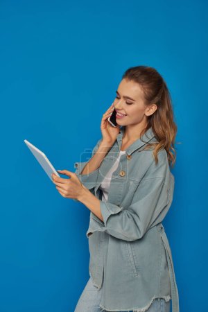 glückliche junge Frau im Gespräch auf Smartphone und mit digitalem Tablet auf blauem Hintergrund, Konnektivität