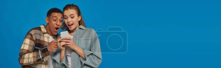 Téléchargez les photos : Couple interracial étonné regardant smartphone sur fond bleu, utilisateurs des médias sociaux, bannière - en image libre de droit