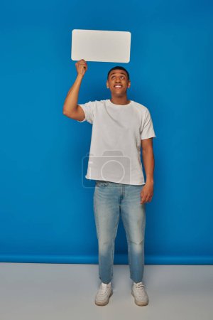 happy african american man in denim jeans holding speech bubble on blue background, full length