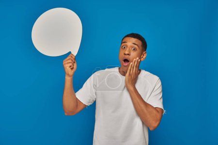hombre afroamericano aturdido con la boca abierta sosteniendo burbuja de habla en blanco sobre fondo azul