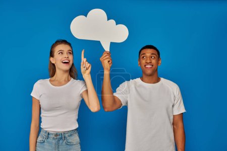 Téléchargez les photos : Heureux couple multiculturel souriant et pointant avec les doigts à bulle de pensée sur fond bleu - en image libre de droit