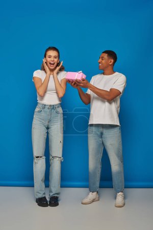 happy african american man presenting wrapped gift to excited girlfriend on blue backdrop