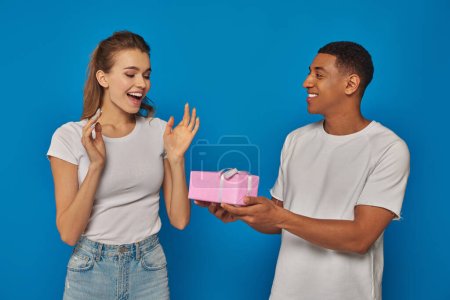 fröhlicher afrikanisch-amerikanischer Mann überreicht verpacktes Geschenk an aufgeregte Freundin vor blauem Hintergrund