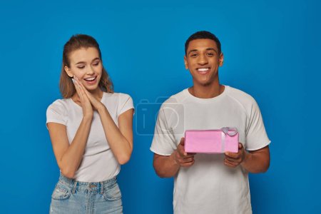 joyeux homme afro-américain tenant cadeau enveloppé près de petite amie excitée sur fond bleu