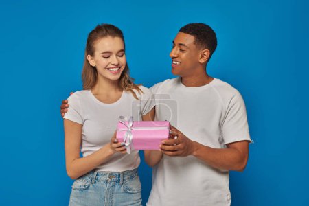 freudiger afrikanisch-amerikanischer Mann überreicht verpacktes Geschenk an aufgeregte Freundin vor blauem Hintergrund
