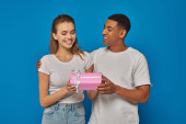 joyful african american man presenting wrapped gift to excited girlfriend on blue backdrop t-shirt #671415154
