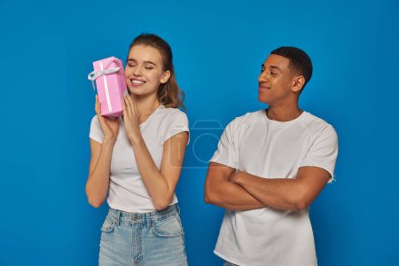 pleased african american man looking at happy girlfriend holding wrapped  gift box on blue backdrop