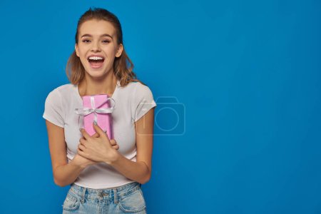 aufgeregte junge Frau hält Geschenkbox in der Hand und blickt in die Kamera auf blauem Hintergrund, festliche Anlässe