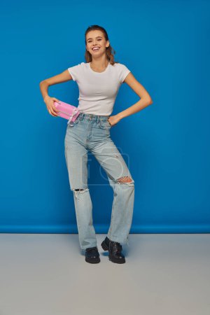 happy young woman in jeans holding gift and looking at camera on blue background, festive occasions