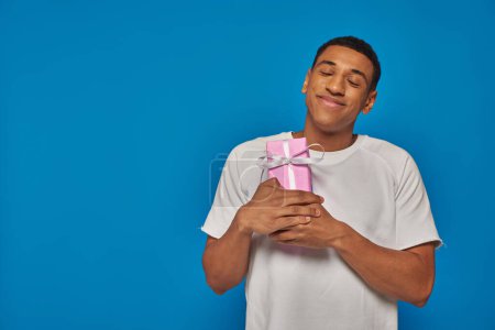 joyous african american man smiling and holding wrapped gift on blue background, festive occasions