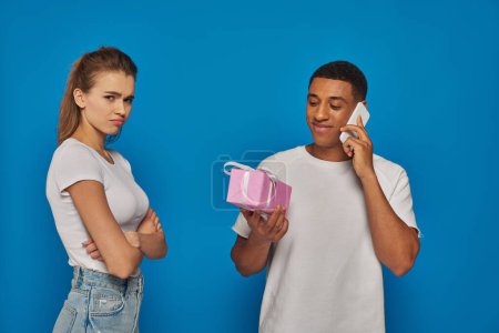 glücklich afrikanisch-amerikanischer Mann telefoniert und hält Geschenk neben beleidigter Frau auf blauem Hintergrund