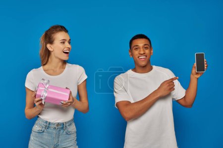Téléchargez les photos : E-commerce, heureux homme afro-américain pointant vers smartphone près de la femme avec un cadeau sur fond bleu - en image libre de droit