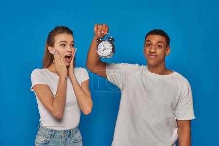 morning time, happy african american man holding alarm clock near astonished woman on blue backdrop