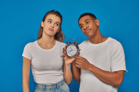 morning time, two displeased interracial couple holding retro alarm clock on blue backdrop