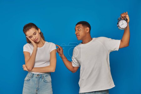 Photo for Awaken african american man holding alarm clock and waking up sleepy woman on blue backdrop - Royalty Free Image