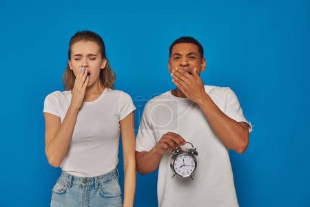 african american man holding alarm clock and yawning with woman on blue backdrop, sleepy couple